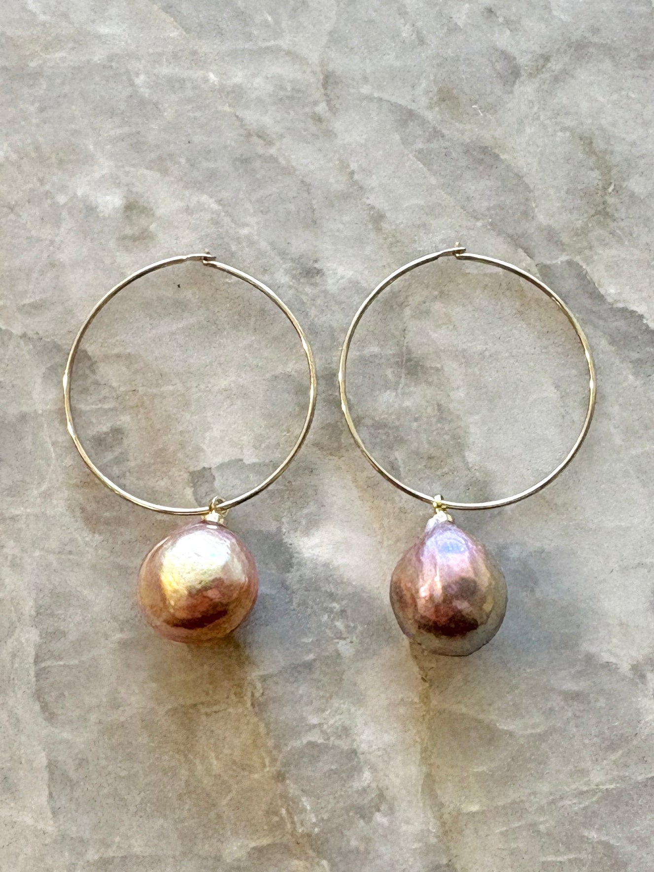 A pair of rose colored fresh water pearl earrings on a thin gold hoop sit on a marble background