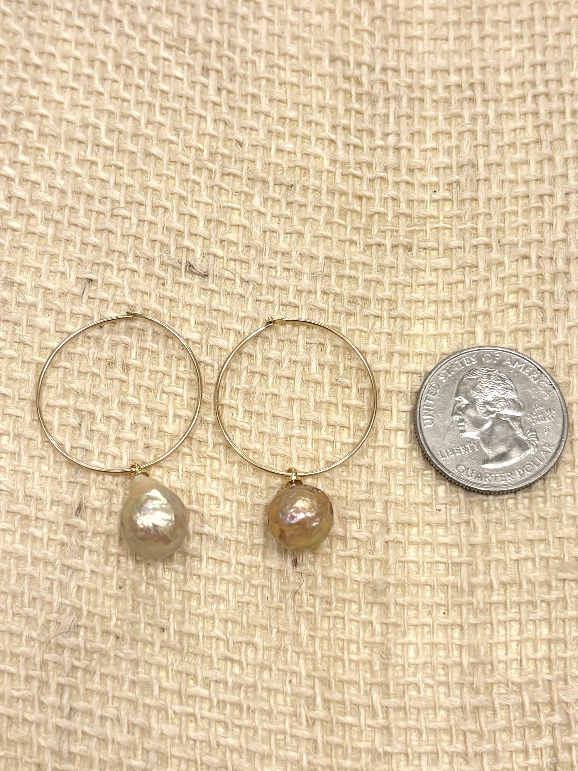 A pair of rose colored freshwater pearls on a thin gold hoop sit on a burlap background next to a quarter for scale. The hoop is approximately the size of the quarter