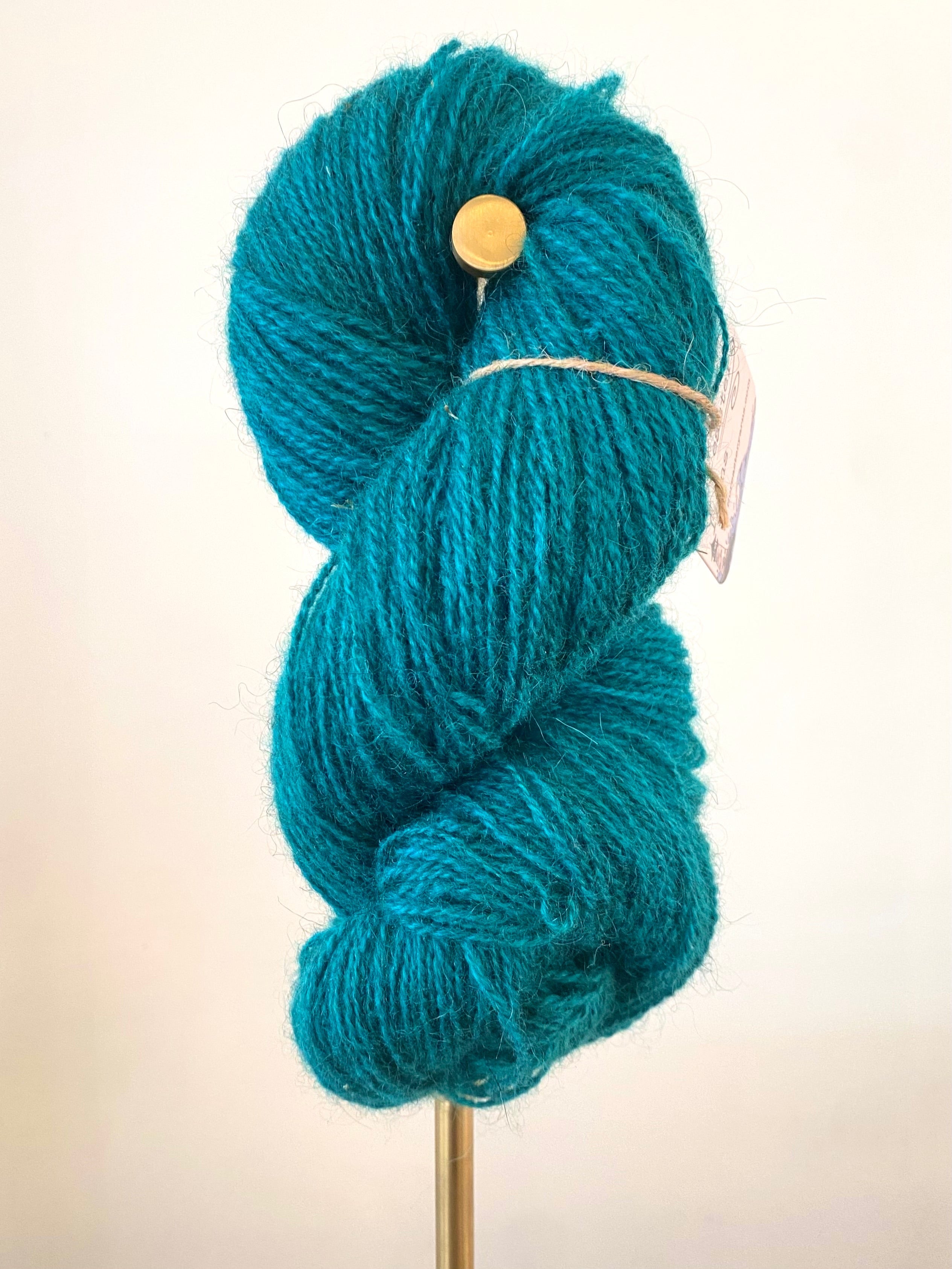 A single skein of teal colored yarn on a gold stand, in front of a white wall. A white tag hangs off the yarn, secured with brown string. The tag has the yarn information