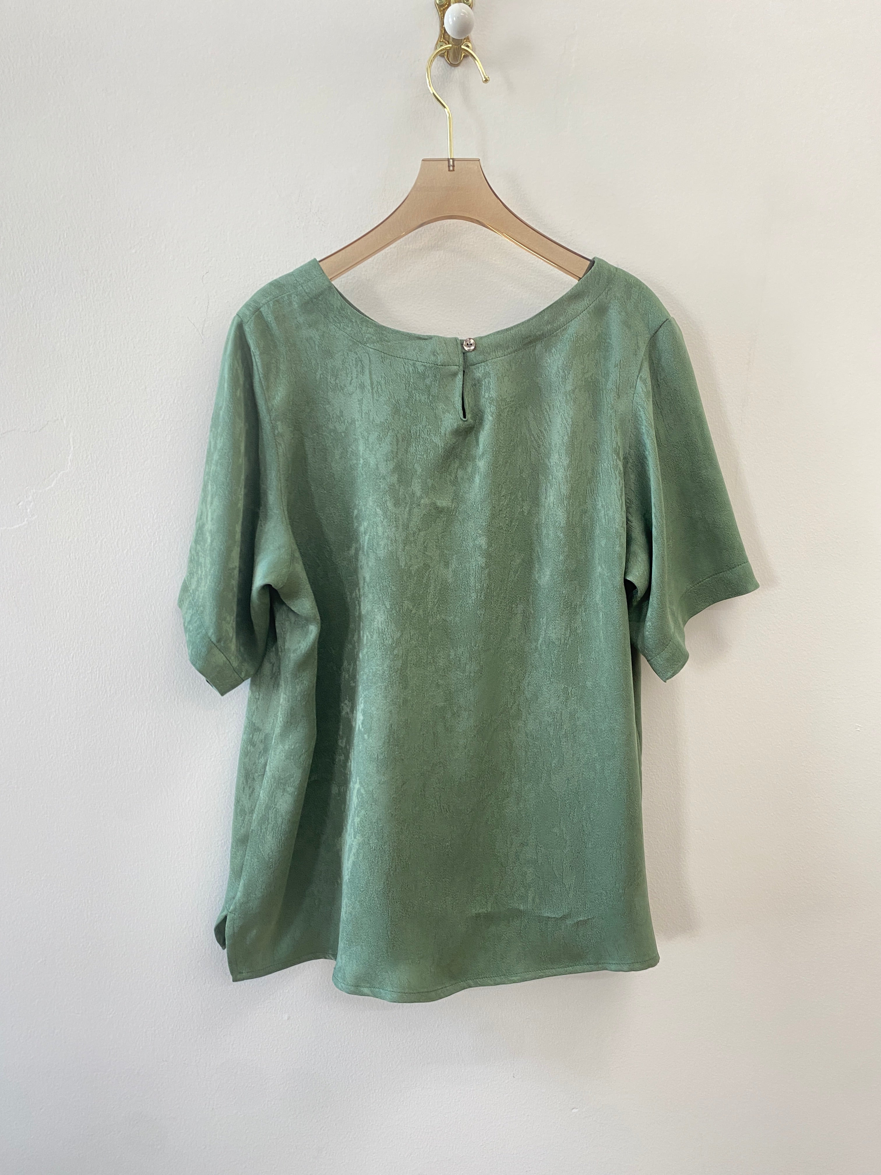 The back of the Jade green top, the button close neckline is visible. The shirt hangs on a brown hanger on a white wall