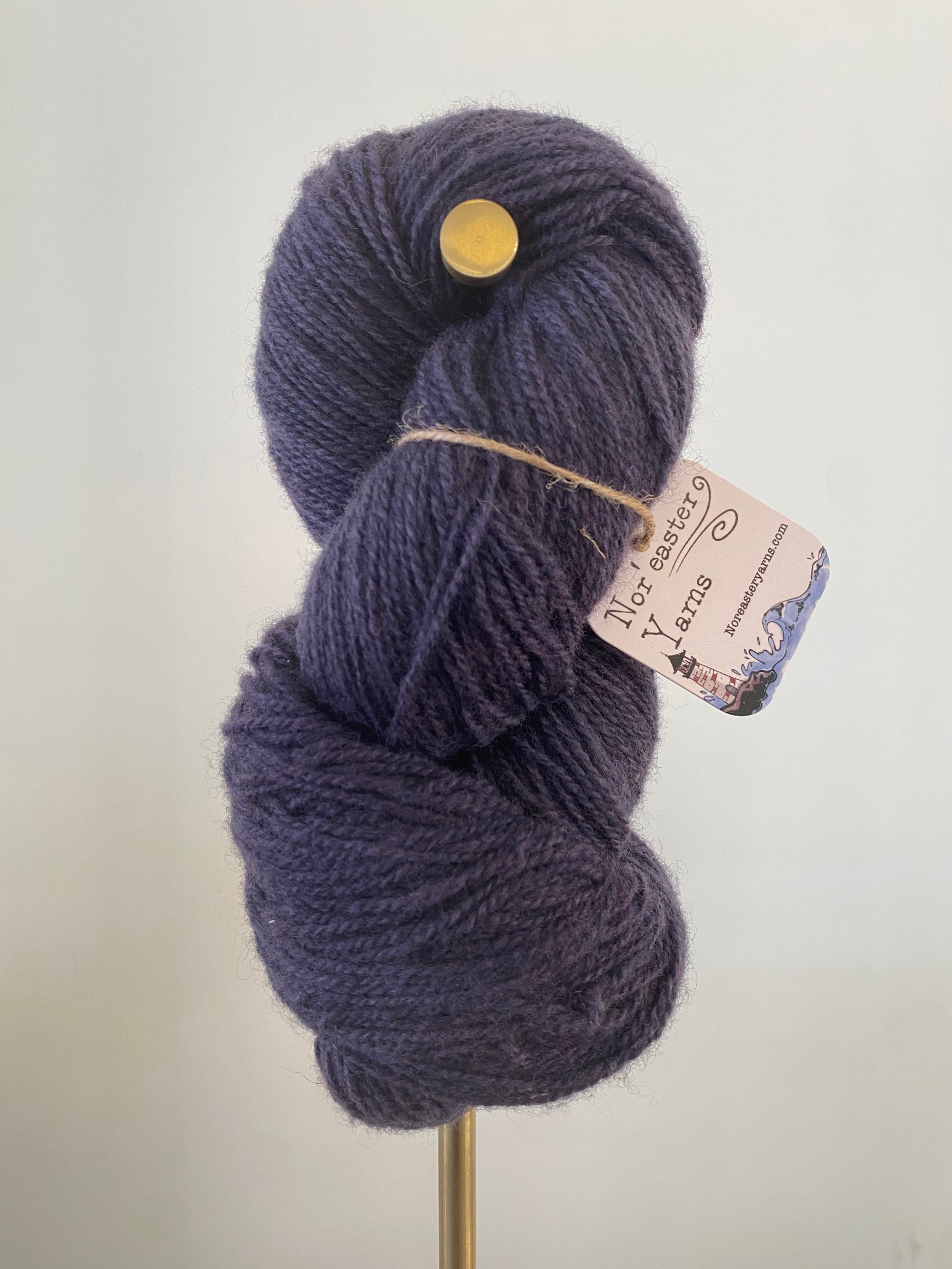 A single skein of dark purple colored yarn on a gold stand, in front of a white wall. A pink hued tag hangs off the yarn, secured with brown string. The tag says Nor’Easter Yarns