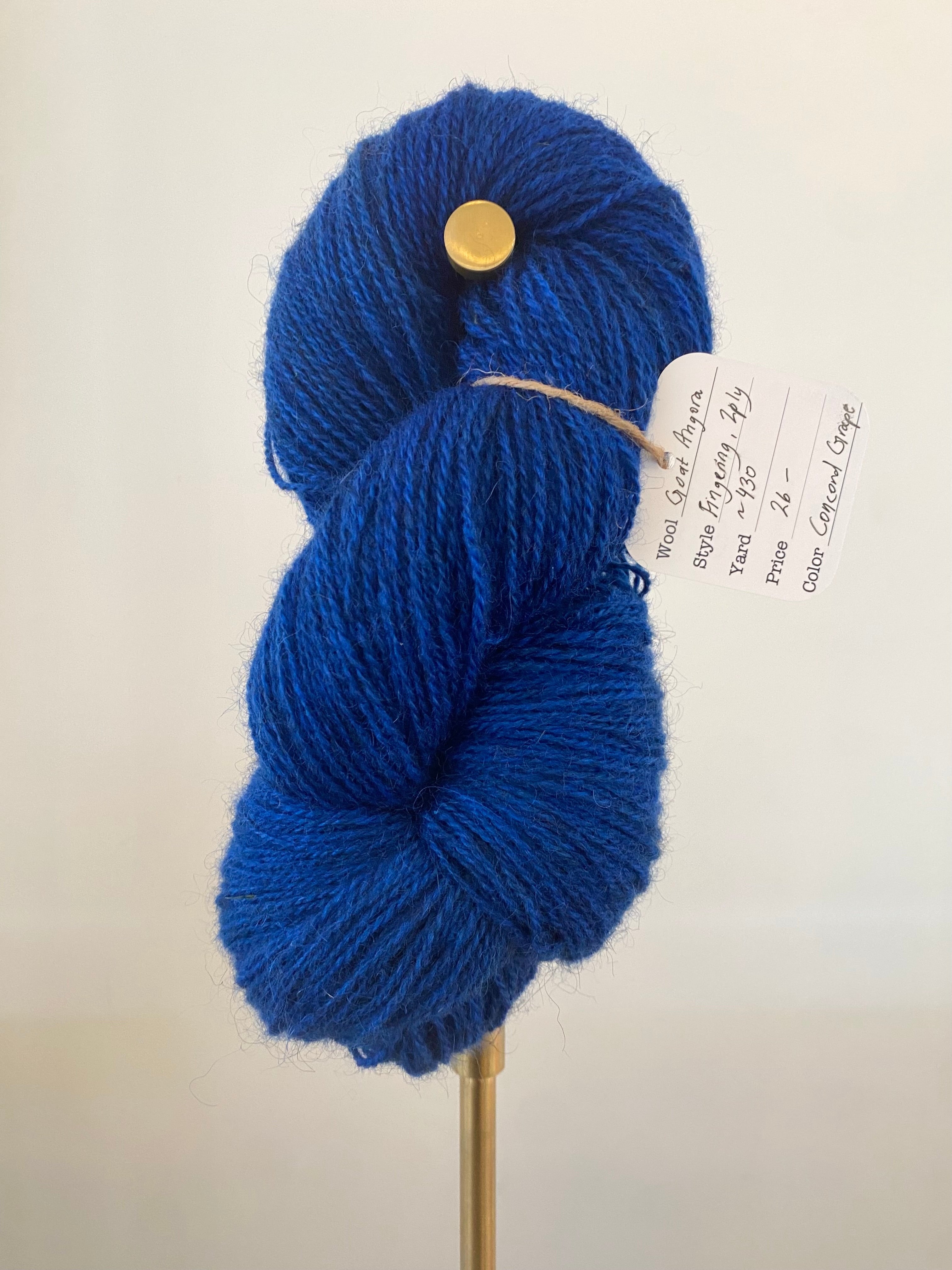 A single skein of blue colored yarn on a gold stand, in front of a white wall. A white tag hangs off the yarn, secured with brown string. The tag has the yarn information