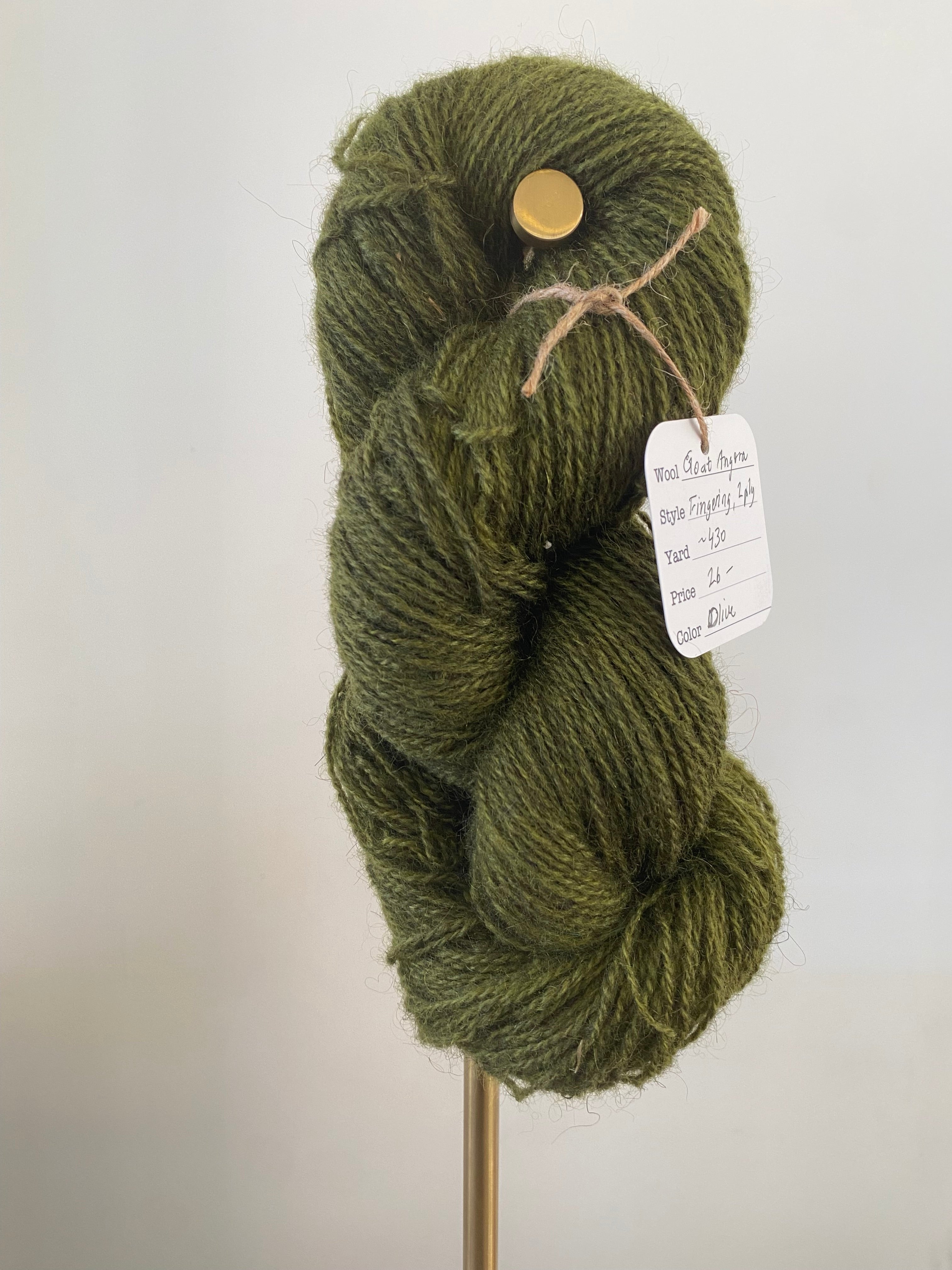 A single skein of olive colored yarn on a gold stand, in front of a white wall. A white tag hangs off the yarn, secured with brown string. The tag has the yarn information