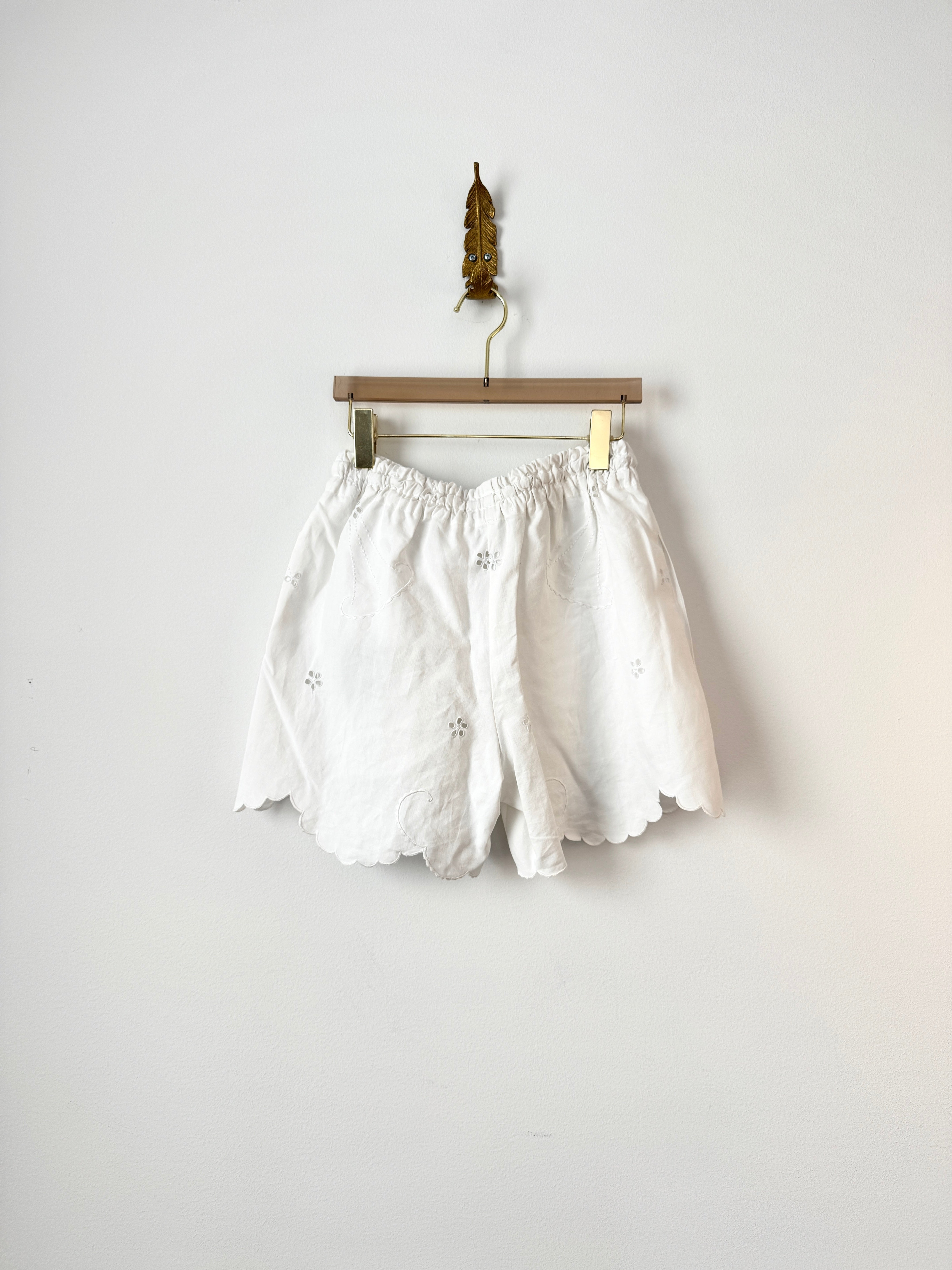White embroidered shorts with a scalloped hem, hanging from a wooden hanger with gold clips against a plain white wall.