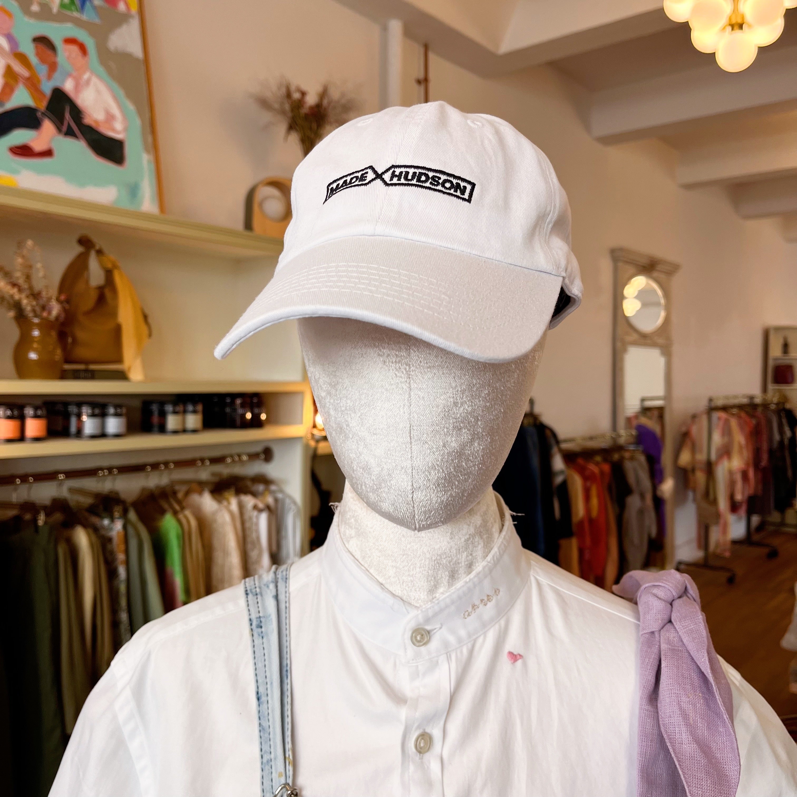 A faceless mannequin in a store wears a white curved brim baseball cap with “Made X Hudson” embroidered on the front.
The mannequin is in a white shirt with a blue strap over one shoulder, and a light purple strap over the other.
Store racks are in the background