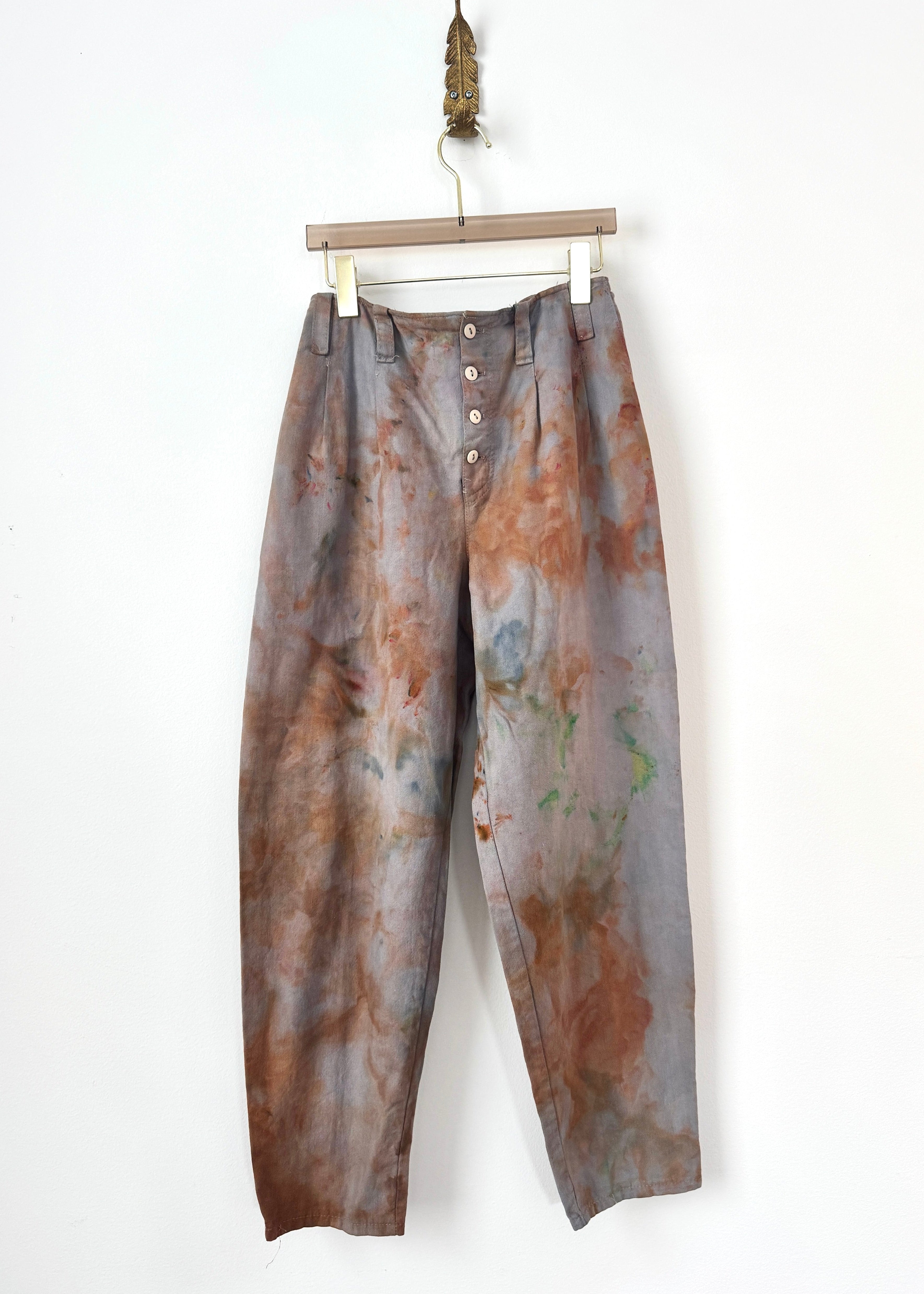 A pair of high-waisted, wide-leg trousers with a button fly, featuring an abstract marbled pattern in brown, gray, and muted green, hanging from a wooden hanger against a plain white wall.