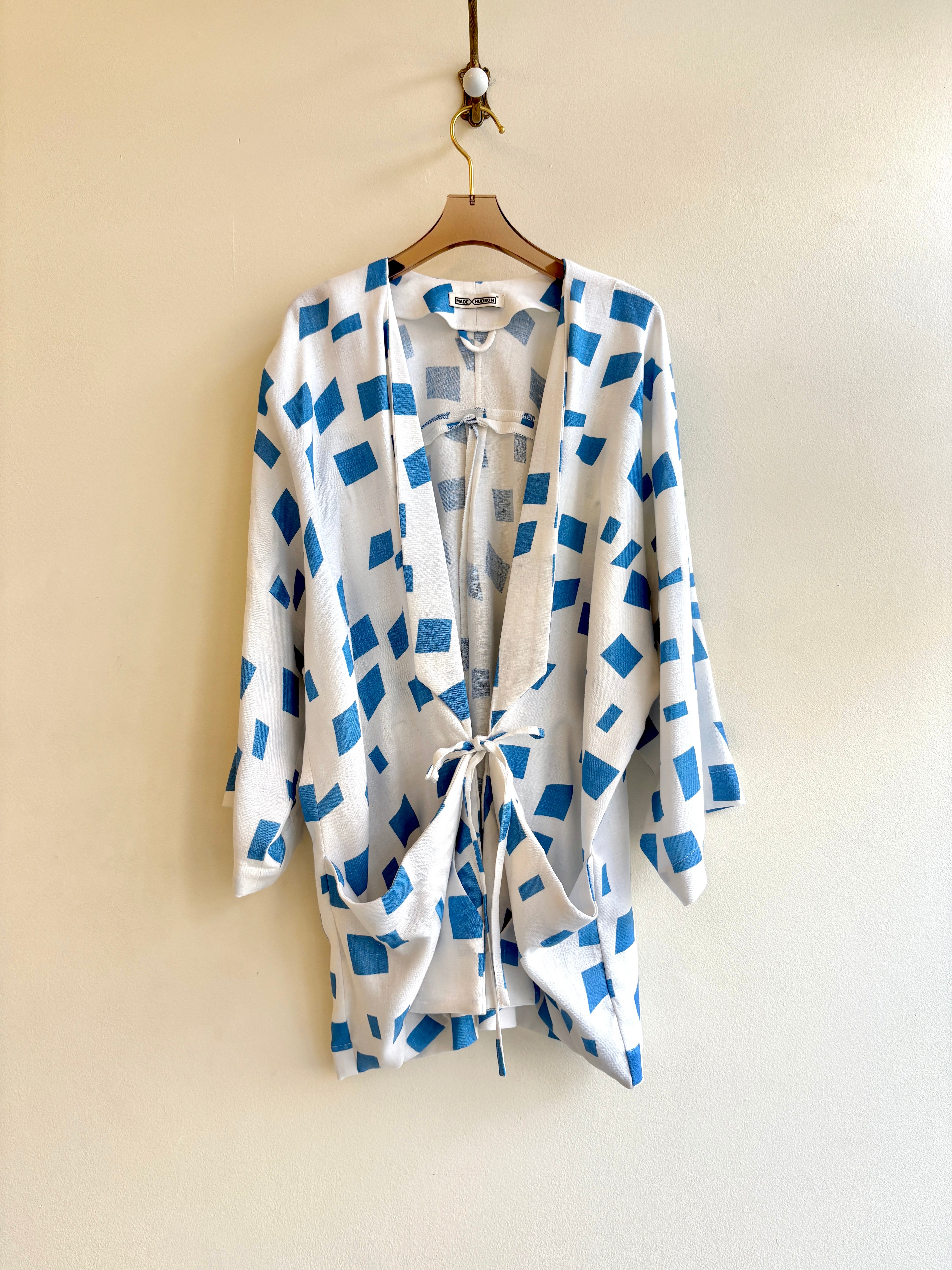 A white with blue squares jacket. The jacket has folded lapels, a tie closure tied in a bow, and two front pockets. The jacket has 3/4 sleeves