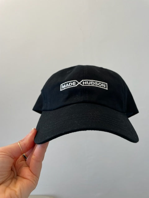 A hand holds a black curved brim baseball hat with “Made X Hudson” embroidered in white on the front.