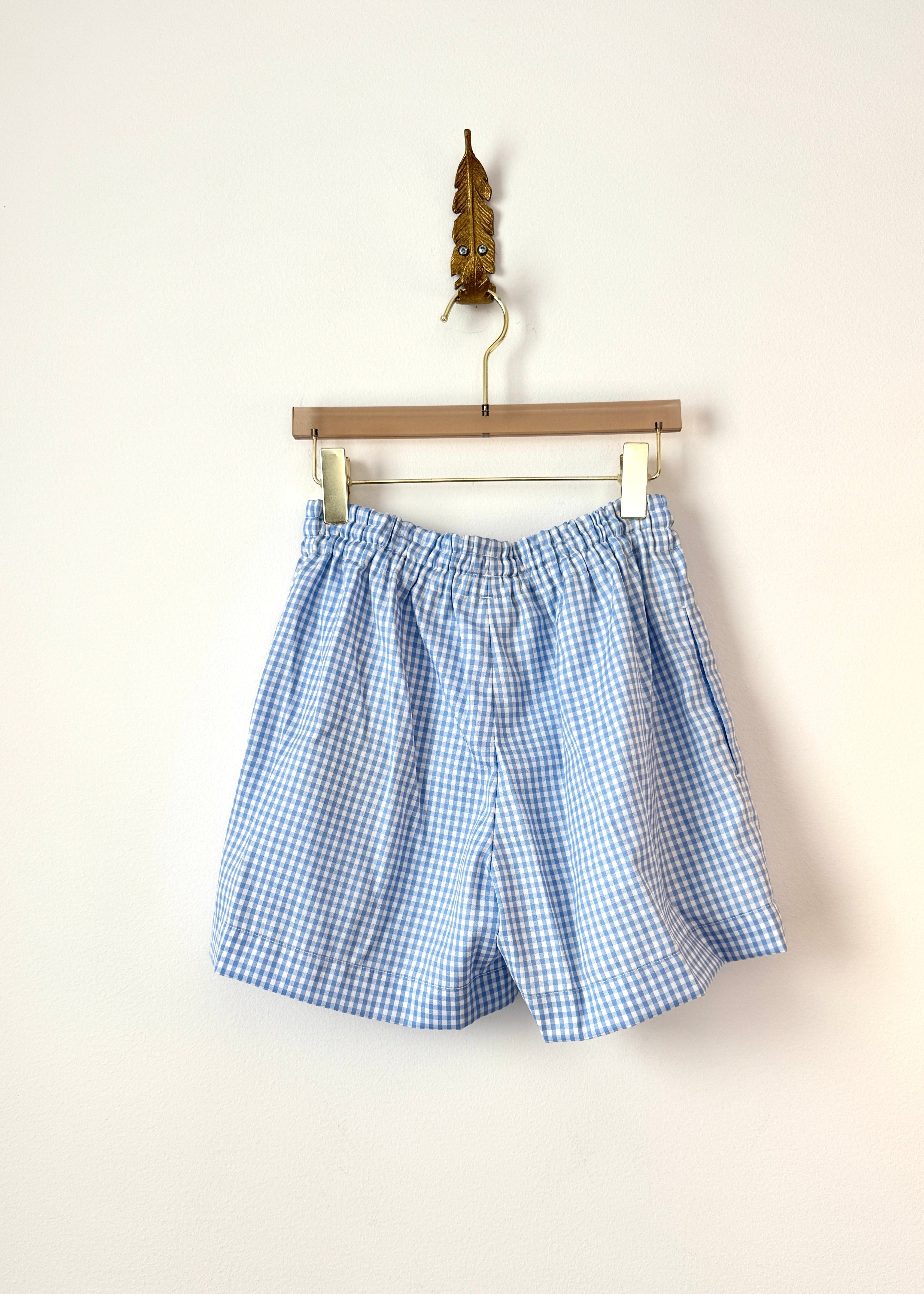 Light blue and white gingham shorts hanging from a wooden hanger against a plain off-white wall.