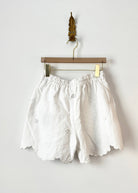 White embroidered shorts with a scalloped hem, hanging from a wooden hanger with gold clips against a plain white wall.